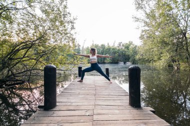 Spor giyimli genç bir kadın doğada yoga yapıyor. O, gölün yanındaki tahta bir platformda yalınayak.