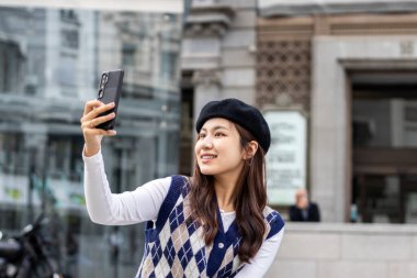 Avrupa 'da bir kentteki bankta oturmuş akıllı telefon kullanan ve selfie çeken Asyalı genç kadın.