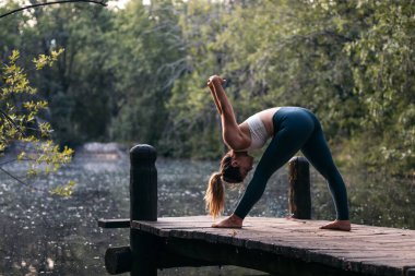 Spor giyimli genç bir kadın doğada yoga yapıyor. O, gölün yanındaki tahta bir platformda yalınayak..