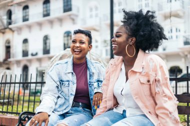 İki siyahi kadın arkadaş kenarda oturmuş gülüyor ve eğleniyorlar.