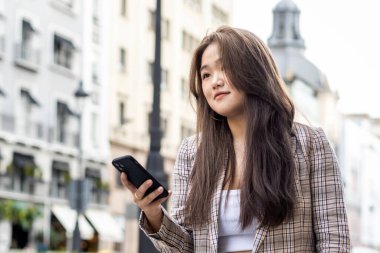 Madrid caddesinde yürüyen Asyalı genç kadın akıllı telefona bakıyor.