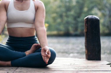 Spor giyim sektöründe meditasyon yapan ve yoga yapan genç bir kadın. O, gölün yanındaki tahta bir platformda yalınayak.
