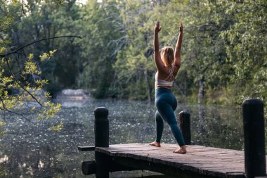 Spor giyimli genç bir kadın doğada yoga yapıyor. O, gölün yanındaki tahta bir platformda yalınayak..
