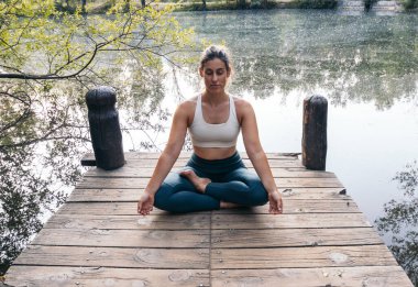 Spor giyim sektöründe meditasyon yapan ve yoga yapan genç bir kadın. O, gölün yanındaki tahta bir platformda yalınayak.