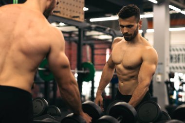 güçlü bir adam aynanın önünde duruyor spor salonunda dambıllar alıyor.