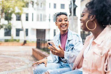 Akıllı telefon kullanan ve konuşan iki siyah kadın arkadaş bir bankta oturup gülüyor ve şehirde eğleniyorlar.