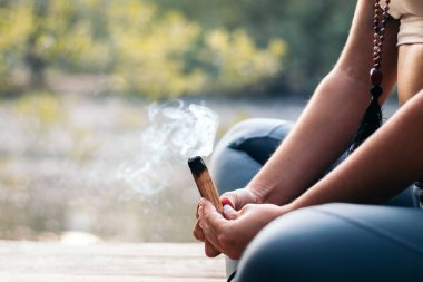 Spor kıyafetleriyle meditasyon yapan ve doğada yoga yapan tanınmamış bir kadına yaklaş. Ahşap bir santo yakıyor..