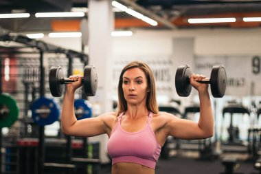 Kadın sporcu spor salonunda aynanın önünde dambıllarla antrenman yapıyor.