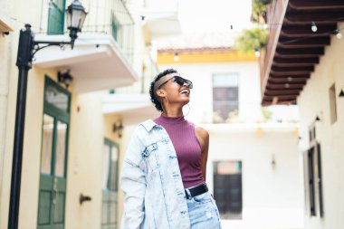 Kısa kıvırcık saçlı, kocaman küpeli havalı bir kadın sokaklarda yürüyor. Günlük kıyafetler ve güneş gözlüğü takıyor.