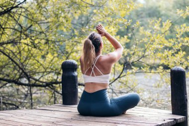 Spor giyim sektöründe meditasyon yapan ve yoga yapan genç bir kadın. O, gölün yanındaki tahta bir platformda yalınayak.