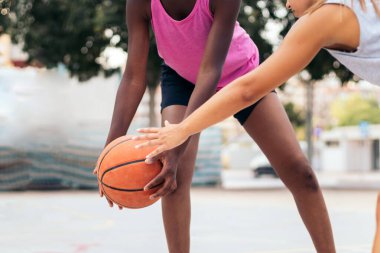 Genç bayan sporcular şehirdeki bir sahada spor kıyafetleriyle basketbol oynuyorlar.