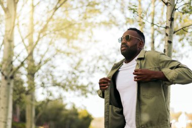 Güneş gözlüklü şık Afro-Amerikan genç adam parkta yürüyor.