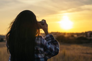 Kareli gömlekli genç bir kadın gün batımında tarlada analog bir kamerayla fotoğraf çekiyor. Gökyüzü turuncu ve görüntüdeki renkler sükuneti iletiyor..