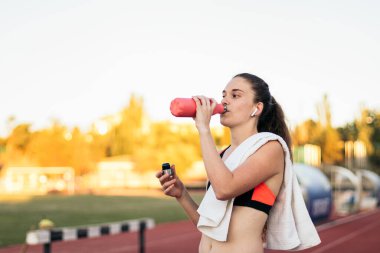 Atletik bir kadın, atletizm eğitiminden sonra kulaklıkla müzik dinliyor ve içme suyu içiyor..