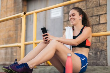 Atletik bir kadın akıllı telefonunu kullanıyor ve atletizm eğitiminden sonra kulaklıkla müzik dinliyor..