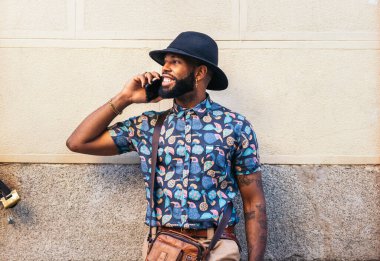 Şapkalı ve alternatif kıyafetli şık siyahi adam telefonda konuşuyor. Şehir sokaklarında duvara yaslanıyor.