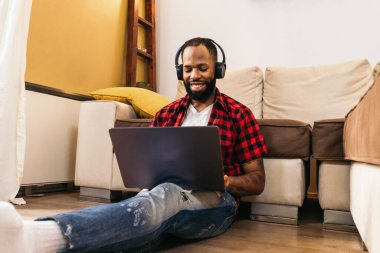 Evde dizüstü bilgisayar kullanan sıradan giysili Afro-Amerikan bir adam. O, kulaklıkla müzik dinlerken oturma odasındaki kanepeye yaslanmış yerde oturuyor. Kişisel bir internet projesine odaklanmış durumda.