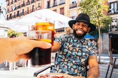 Süslü, boyalı tırnaklı, şapkalı, içki içen ve kadeh kaldıran siyahi bir adam Madrid şehrinde bir yaz gününde terasta oturuyor.