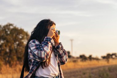 Kareli gömlekli ve günlük kıyafetlerle gün batımında tarlada analog bir kamerayla fotoğraf çeken genç bir kadın. Görüntü, retro ve vintage konseptini iletir