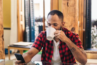 Sabahları işten önce evde kahve içerken akıllı telefon kullanan sıradan giyinmiş Afro-Amerikalı bir adam.