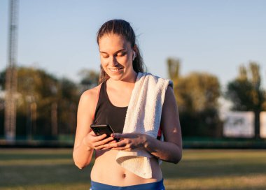 Atletik bir kadın akıllı telefonunu kullanıyor ve atletizm eğitiminden sonra kulaklıkla müzik dinliyor..
