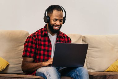 Laptop bilgisayar ve kulaklıklarla uzaktan çalışan gülümseyen siyah bir adam. Telekomünikasyon ve ev ofisi kavramı.