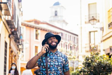 Alternatif hippi görünümlü şık siyahi bir adam güneşli bir yaz gününde telefonda konuşurken bir şehrin sokaklarında yürüyor.