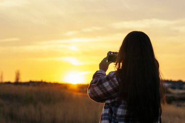 Kareli gömlekli genç bir kadın gün batımında tarlada analog bir kamerayla fotoğraf çekiyor. Gökyüzü turuncu ve görüntüdeki renkler sükuneti iletiyor..