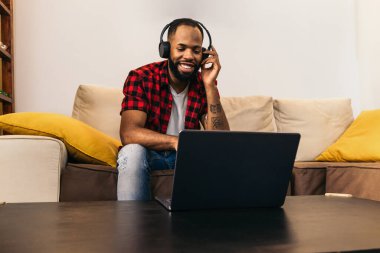 Günlük kıyafetleri ve kulaklıkları olan siyah bir adam kanepenin üzerinde dizüstü bilgisayarın önünde oturuyor. Görüntü, bilgisayar üzerinden film ve video izleme kavramının yanı sıra evdeki tele-çalışma ve video görüşmelerini de aktarıyor..