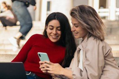 Üniversitenin merdivenlerinde dizüstü bilgisayar ve cep telefonuyla oturan iki öğrenci kız. Yakın plan.