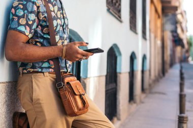 Alternatif kıyafetli şık siyah adam şehrin sokaklarında duvara dayanan akıllı telefon kullanıyor.