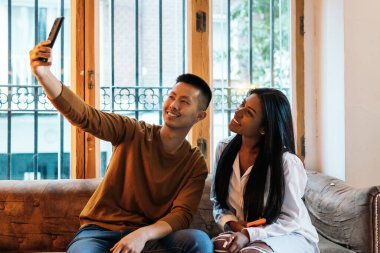 Çok ırklı arkadaşlar, geniş pencereli bir koltukta selfie çekiyorlar. Akıllı telefon kamerasına bakıp gülümsüyorlar.