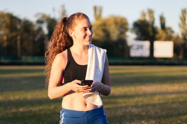 Atletik bir kadın akıllı telefonunu kullanıyor ve atletizm eğitiminden sonra kulaklıkla müzik dinliyor..