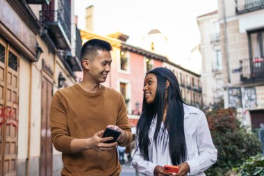 Asyalı erkek ve Latin Amerikalı kadın bir Avrupa şehrinin caddelerinde yürürken konuşuyorlar ve akıllı telefon kullanırken iyi vakit geçiriyorlar.