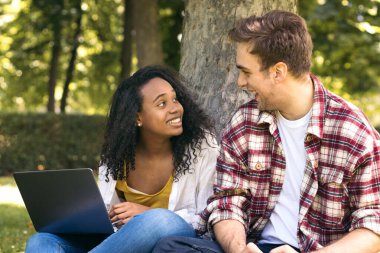 Parkta dizüstü bilgisayar kullanarak oturan iki çok ırklı genç öğrenci. Birlikte çalışıp eğleniyorlar.
