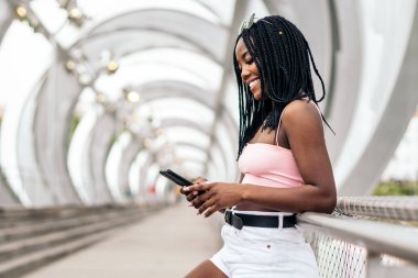 Renkli yaz kıyafetleri içinde Afro-Amerikan bir kızın portresi. Akıllı telefonu kullanırken şehir köprüsüne yaslanıyor.