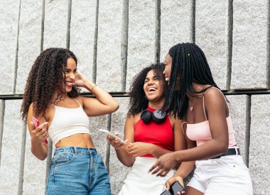 Üç Afro saçlı Afro-Amerikalı kız gülüyor ve akıllı telefon kullanarak şehirde iyi vakit geçiriyorlar.