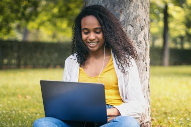 Sarı tişörtlü Afro-Amerikalı kız kampüs çimlerinde oturmuş dizüstü bilgisayarla üniversite ödevi yapıyor. Kulaklıkla müzik dinleyip ders çalışıyor gibi görünüyor.