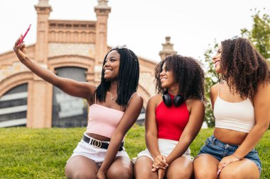 Parkta oturan üç Afrikalı Amerikalı kız iyi vakit geçiriyorlar. Akıllı telefon kullanıyorlar ve sosyal ağlardan resimler ve videolar görüyorlar.