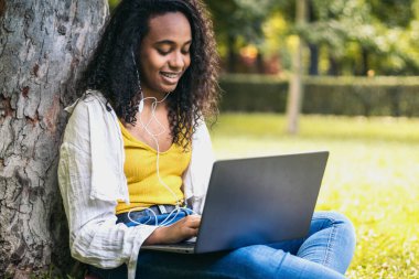 Sarı tişörtlü Afro-Amerikalı kız kampüs çimlerinde oturmuş dizüstü bilgisayarla üniversite ödevi yapıyor. Kulaklıkla müzik dinleyip ders çalışıyor gibi görünüyor.