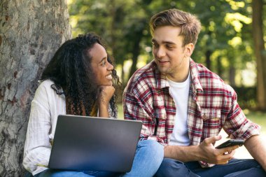 Parkta dizüstü bilgisayar kullanarak oturan iki çok ırklı genç öğrenci. Birlikte çalışıp eğleniyorlar.