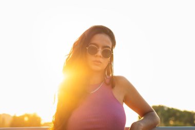 Güneş gözlüklü genç bir Latin kadının gün batımında çekilmiş bir portresi. Kameraya bakıyor ve poz veriyor.