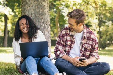Parkta dizüstü bilgisayar ve akıllı telefon kullanarak oturan iki çok ırklı genç öğrenci. Birlikte çalışıp eğleniyorlar.