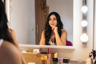 Neşeli genç gelin, makyaj masasında aynada kendine bakıyor. Düğünü için mutlu ve heyecanlı..