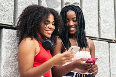 Afro saçlı iki Afrikalı Amerikalı kız parkta akıllı telefon kullanarak gülüyorlar.