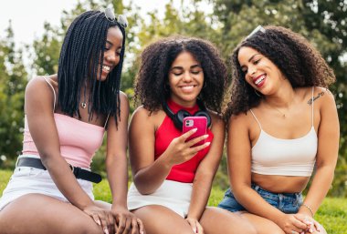 Parkta oturan üç Afrikalı Amerikalı kız iyi vakit geçiriyorlar. Akıllı telefon kullanıyorlar ve sosyal ağlardan resimler ve videolar görüyorlar.