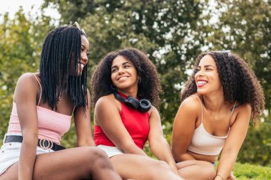 Üç Afro saçlı Afro-Amerikalı kız parkta oturmuş gülüyor ve eğleniyorlar. Yazlık giysiler giyip birlikte vakit geçirmekten hoşlanıyorlar.