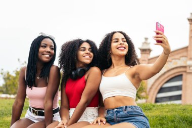 Üç Afro saçlı Afro-Amerikalı kız parkta oturmuş gülüyor, eğleniyor ve akıllı telefon kullanıyor.