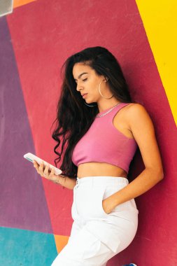 Renkli bir duvara yaslanan ve akıllı telefon kullanan yaz elbiseli Latin bir genç kadının fotoğrafını çek.