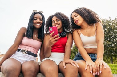 Üç Afro saçlı Afro-Amerikalı kız parkta oturmuş gülüyor, eğleniyor ve akıllı telefon kullanıyor.